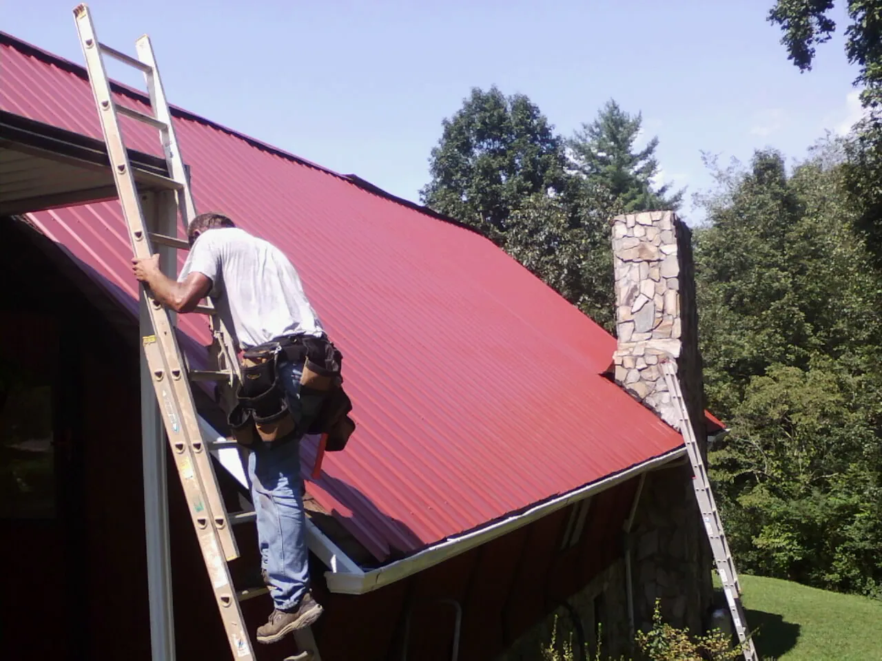 Roofer on a ladder beside a red metal roof and stone chimney — Soffit & Fascia Repair by Roofing Brothers in Lobelville